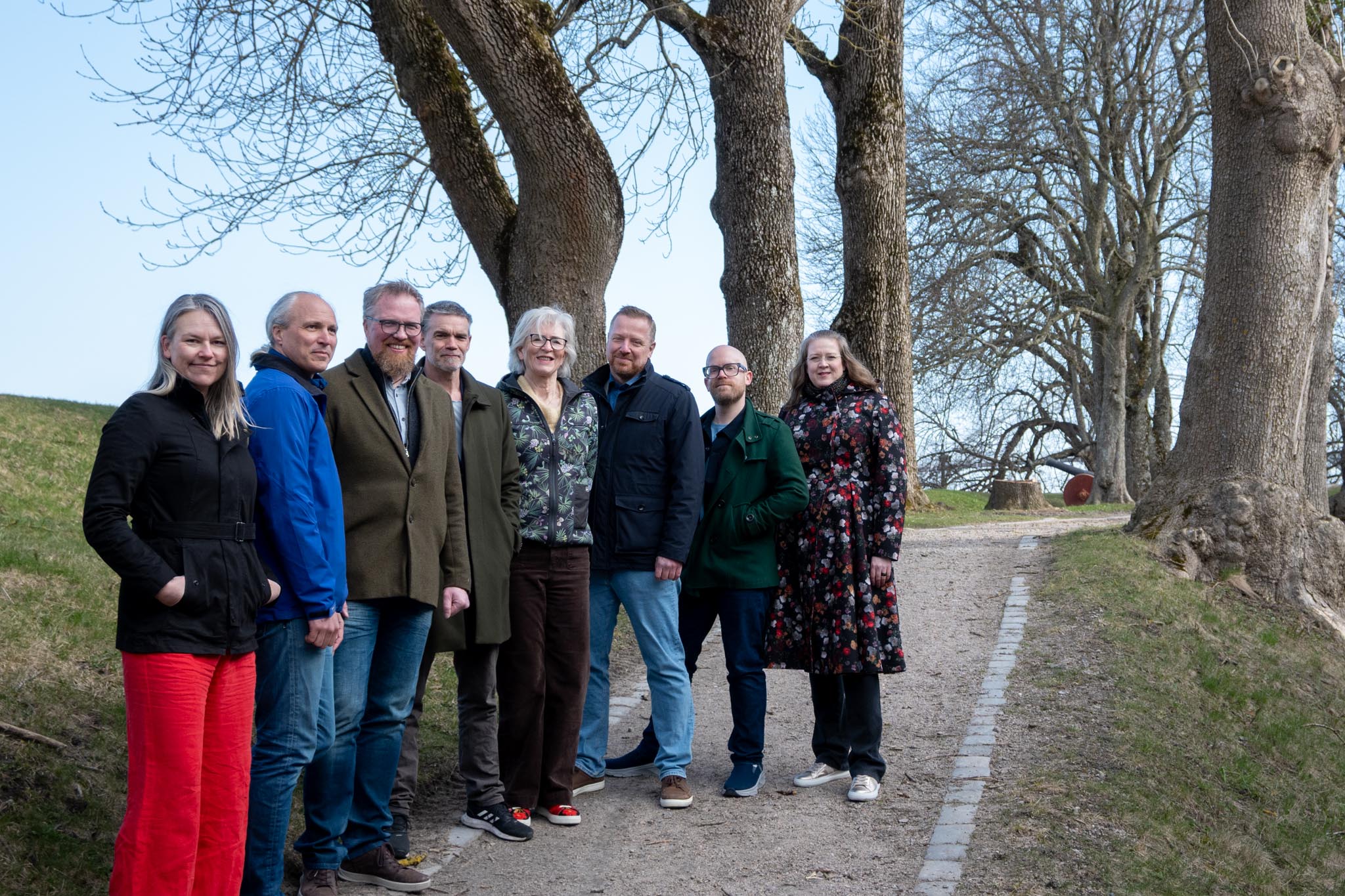 En gruppe mennesker står foran et stort tre. I bakgrunnen ser du blå himmel og andre trær.